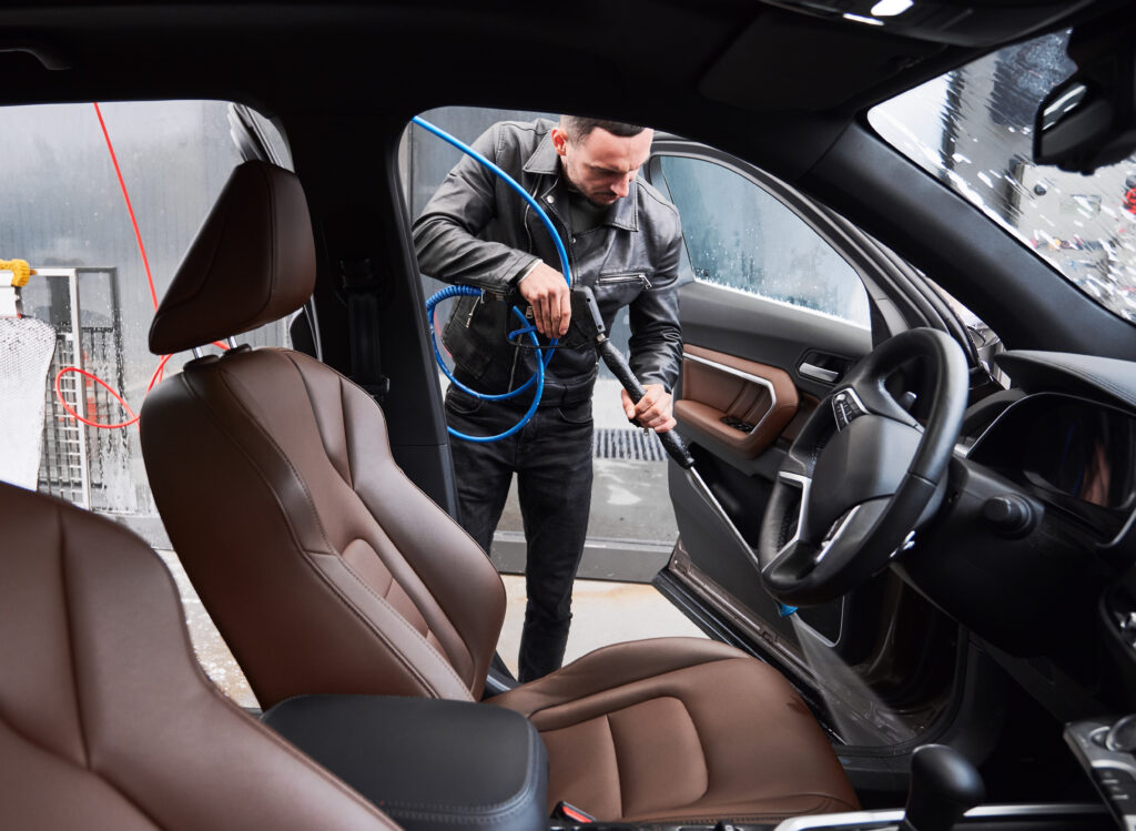 View from inside a car on young man washing car on carwash station outdoor. Handsome driver cleaning automobile, using high pressure water.